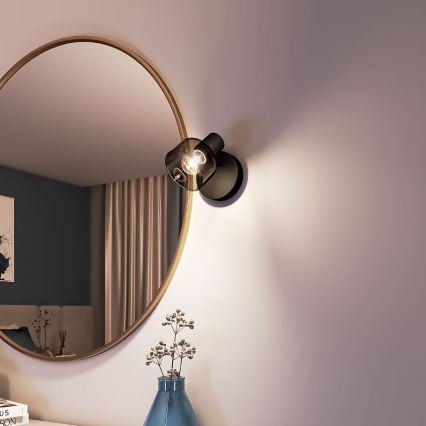 Nástenná lampa svieti v interiéri. Je umiestnená na stene vedľa okrúhleho zrkadla a vázy s kvetmi. Lampa je čierna s priehľadnou sklenenou časťou.