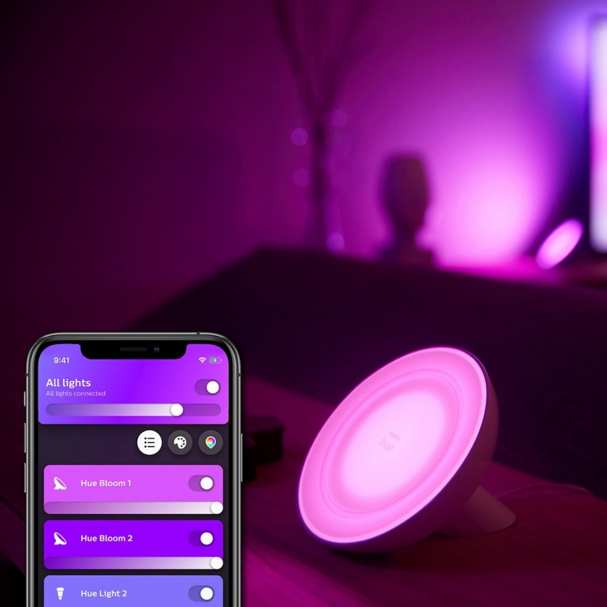 Mobilný telefón zobrazuje aplikáciu na ovládanie osvetlenia. Aplikácia ukazuje zapnuté svetlá „Hue Bloom 1“, „Hue Bloom 2“ a „Hue Light 2“. Na stole je zapnuté kruhové svetlo s názvom „Hue“.
