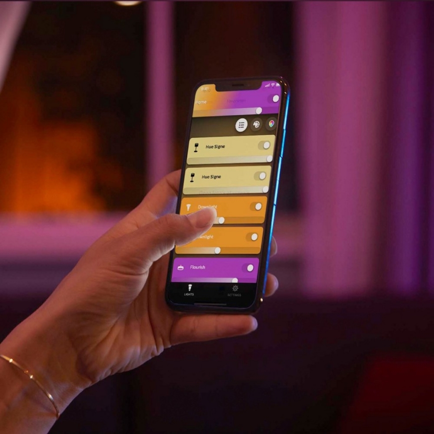 Ruka ovláda smartfón s aplikáciou na ovládanie osvetlenia. Aplikácia zobrazuje prepínače pre „Hue Signe“, „Downlight“, „Twilight“, „Flourish“. Na pozadí je rozostrený interiér s fialovým osvetlením.