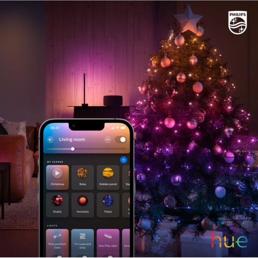 Mobilný telefón zobrazuje aplikáciu na ovládanie svetiel. Aplikácia umožňuje výber rôznych svetelných scén, ako sú Christmas, Ibiza, Golden pond, Osaka, Honolulu a Tokyo. V pozadí je vianočný stromček s farebnými svetielkami a nábytkom v obývacej izbe.