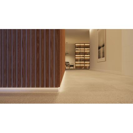 LED pás svieti pozdĺž podlahovej lišty z dreva pri stene. Lišta sa nachádza v chodbe s drevenou stenou a knižnicou v pozadí. Chodba má svetlý povrch podlahy.