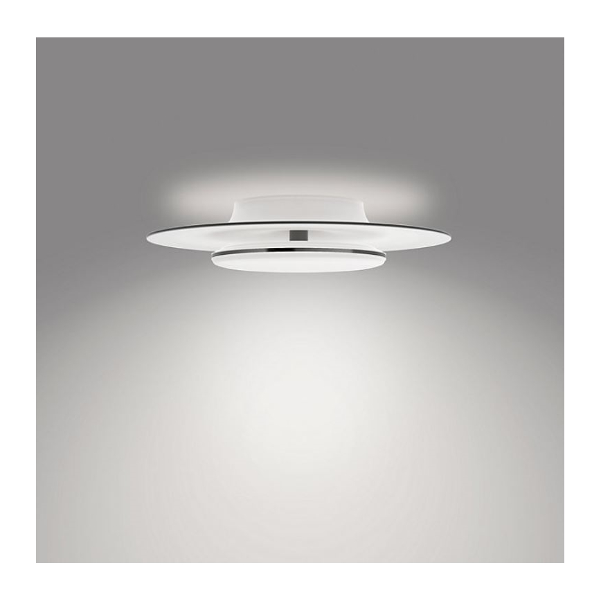 Svietidlo svieti na strop. Je biele s dvoma kruhovými vrstvami a tenkým kovovým prstencom. Je namontované na svetlom sivom pozadí.