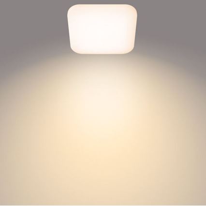 Svietidlo svieti. Je štvorcové, s oblými rohmi, biele a pripevnené k stropu. Svetlo sa šíri po svetložltej stene.