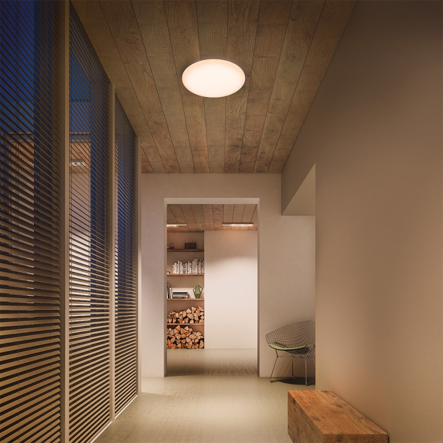 Svetlo svieti v chodbe. Okrúhle stropné svietidlo osvetľuje priestor s dreveným stropom a dvernými krídlami s žalúziami. V pozadí sa nachádza polica s knihami a drevo na kúrenie.