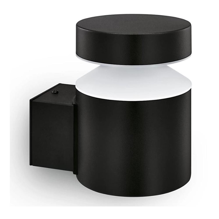 Čierna nástenná lampa svieti. Horná časť lampy je valcovitá s bielym difúzorom. Lampa je pripevnená na stenu štvorcovou základňou.