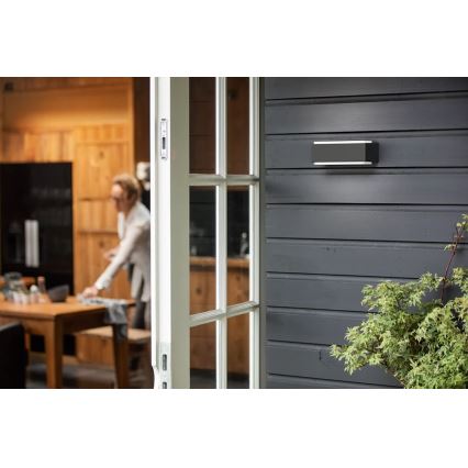 Obdĺžnikové vonkajšie nástenné svietidlo je pripevnené na tmavosivej drevenej stene. Svietidlo je v popredí, za ním je čiastočne viditeľný otvorený dverový rám a rozostrený interiér s kuchyňou a ženou.