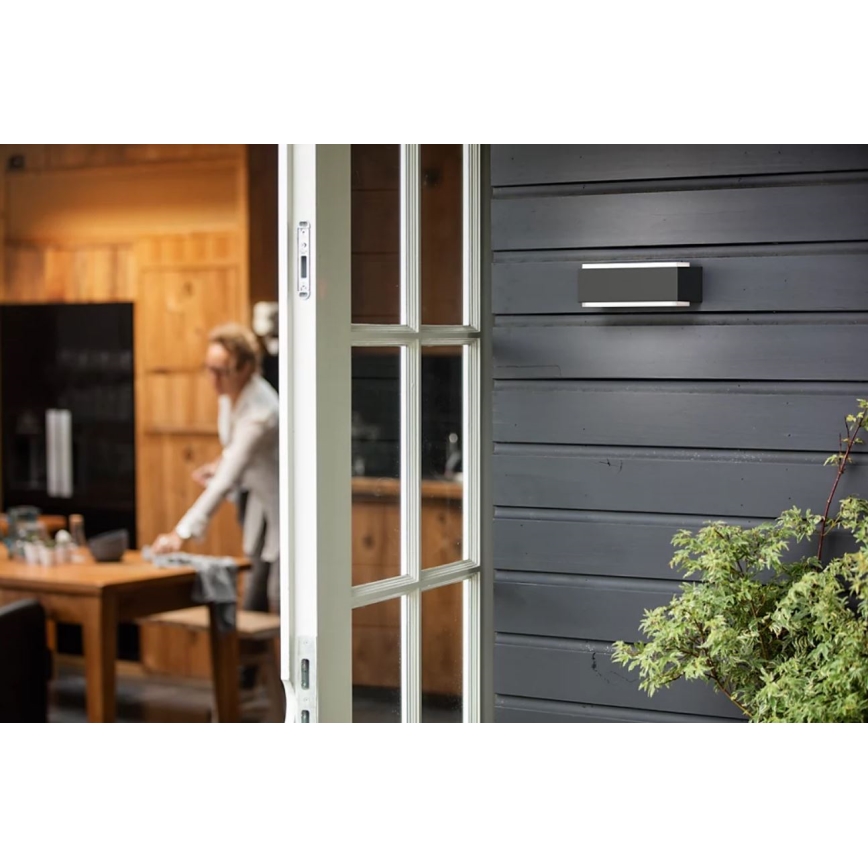 Obdĺžnikové vonkajšie nástenné svietidlo je pripevnené na tmavosivej drevenej stene. Svietidlo je v popredí, za ním je čiastočne viditeľný otvorený dverový rám a rozostrený interiér s kuchyňou a ženou.