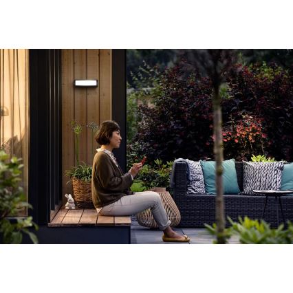 Žena sedí na drevenej lavičke pred domom a drží v ruke mobilný telefón. Na stene domu je namontované vonkajšie LED svietidlo. Okolitý priestor tvorí záhradná terasa s ratanovou sedačkou a zeleňou.