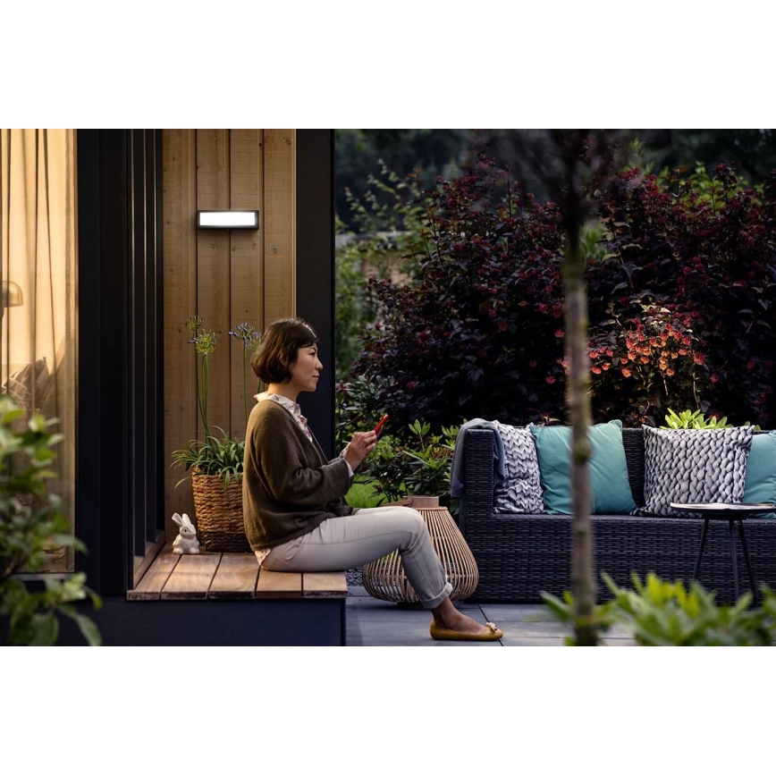 Žena sedí na drevenej lavičke pred domom a drží v ruke mobilný telefón. Na stene domu je namontované vonkajšie LED svietidlo. Okolitý priestor tvorí záhradná terasa s ratanovou sedačkou a zeleňou.