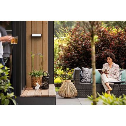 Žena číta knihu na vonkajšej sedačke. Sedačka sa nachádza na terase obklopenej zeleňou a vonkajším osvetlením. Na terase sú aj dekoratívne lampáše a kvetináče.