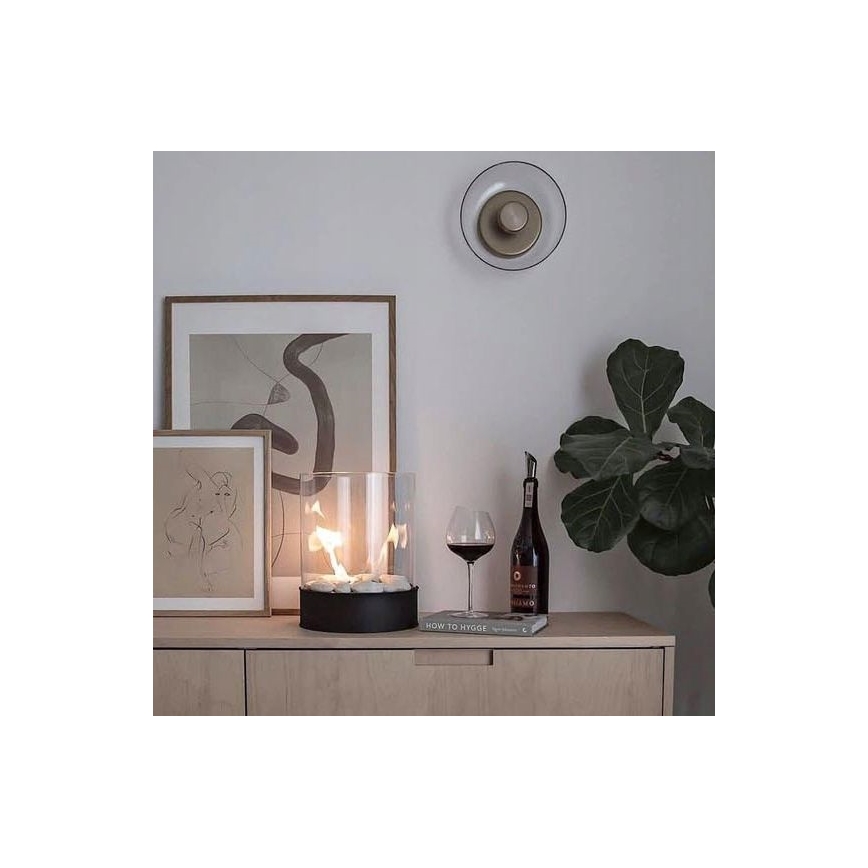 Biokrb horí v sklenenom valci na drevenom nábytku. Vedľa neho je pohár červeného vína, kniha s názvom „HOW TO HYGGE“ a fikus. Na stene visí obraz a nástenné svietidlo.