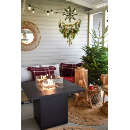 Čtvercový ohňový stôl horí v exteriéri. Nachádza sa na terase s vianočnou výzdobou, vrátane vianočného stromčeka a veniec.