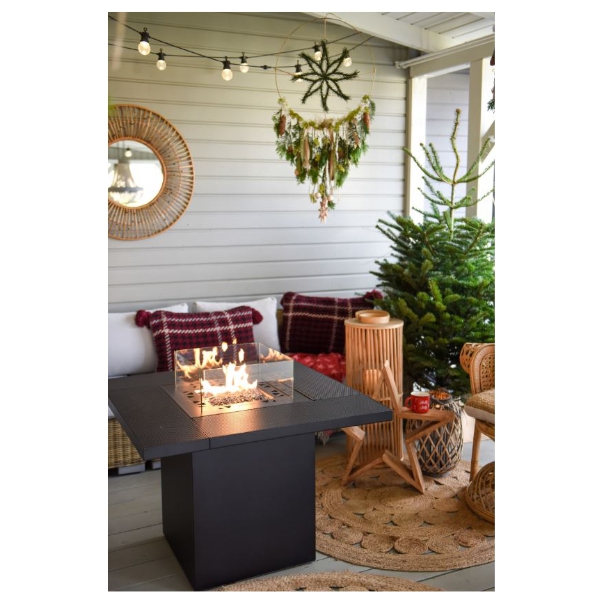 Čtvercový ohňový stôl horí v exteriéri. Nachádza sa na terase s vianočnou výzdobou, vrátane vianočného stromčeka a veniec.