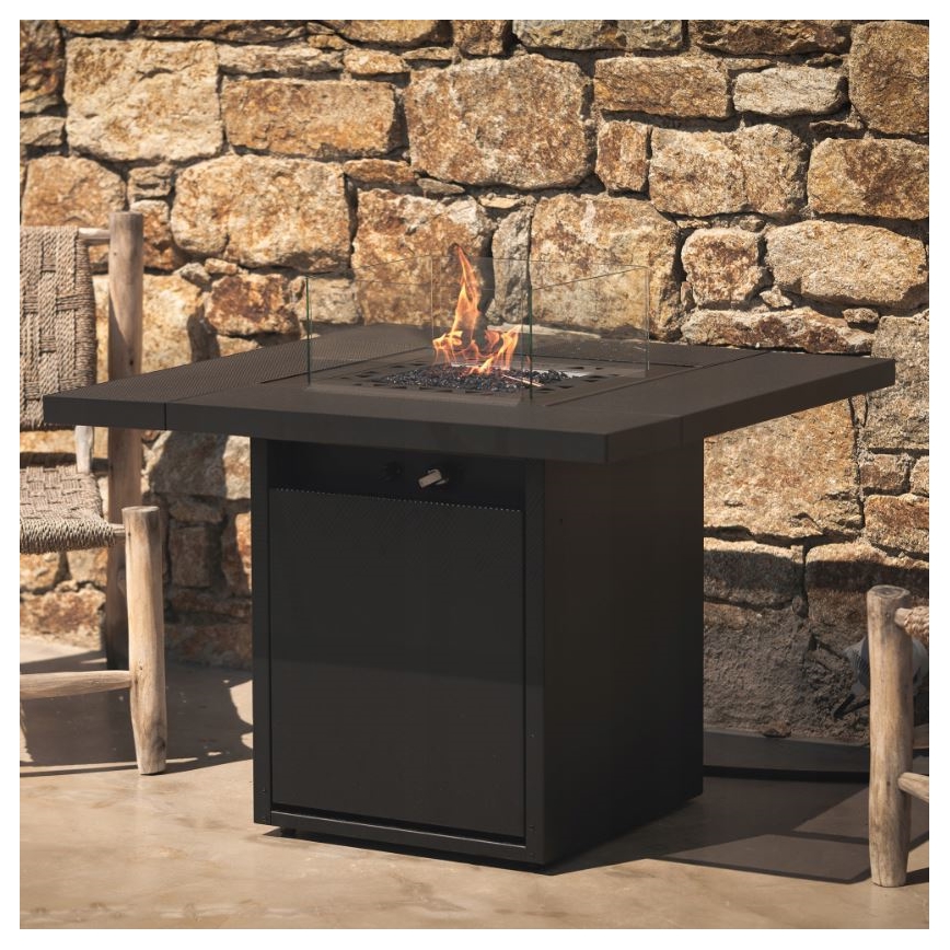 Záhradný krb horí. Má štvorcový tvar s tmavosivým povrchom, sklenený štít a plynový horák. Nachádza sa pred kamennou stenou, vedľa dvoch drevených stoličiek.