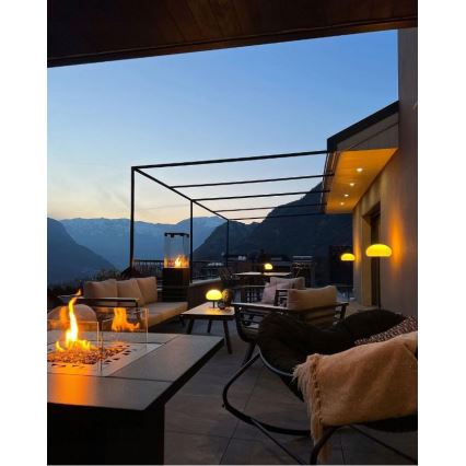 Vonkajší krb horí na terase s výhľadom na hory. Na terase je posedenie s nábytkom, osvetlené lampami. Počasie je zrejme súmračné.