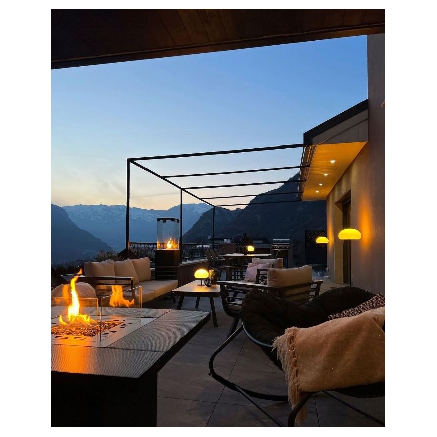 Vonkajší krb horí na terase s výhľadom na hory. Na terase je posedenie s nábytkom, osvetlené lampami. Počasie je zrejme súmračné.