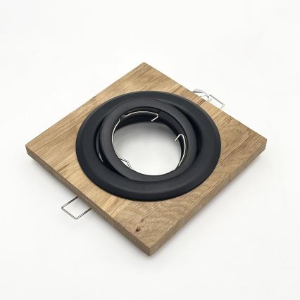 Drevený rámček s dvoma čiernymi kruhovými kovovými prstencami slúži ako držiak pre svietidlo. Je položený na bielom podklade.