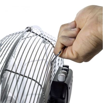 Ruka otvára ochrannú mriežku kovového stojanového ventilátora s čiernou základňou a ovládacími tlačidlami. Ventilátor je na bielom pozadí.
