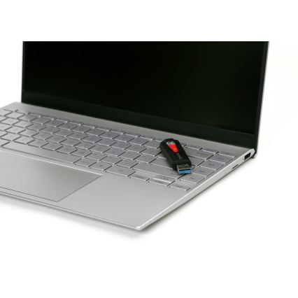 USB kľúč leží na klávesnici strieborného prenosného počítača. Počítač je zatvorený. Kľúč je čierny s červeným detailom.