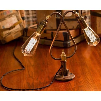 Dvojramenná stolová lampa s Edisonovými žiarovkami svieti na drevenom stole. Lampa je pripevnená k základni pomocou kovového držiaka. Okolím sú starožitné škatule.