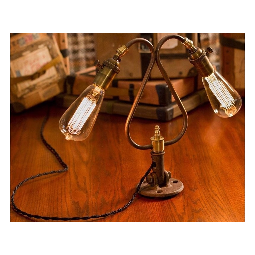 Dvojramenná stolová lampa s Edisonovými žiarovkami svieti na drevenom stole. Lampa je pripevnená k základni pomocou kovového držiaka. Okolím sú starožitné škatule.