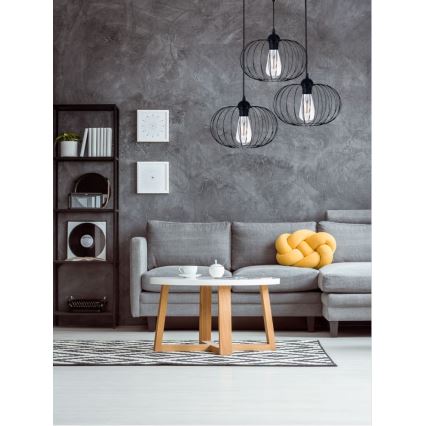 Trojica čiernych kovových závesných lámp s textilnými tienidlami visí nad šedou sedačkou. Lampa je umiestnená v obývacej izbe s dreveným konferenčným stolíkom a policou.