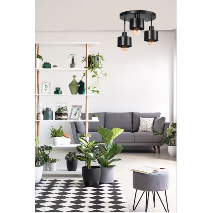 Trojramenné stropné svietidlo svieti v modernej obývacej izbe. Svietidlo je čierne, s cylindrickými tienidlami. Izba obsahuje sivú sedačku, police s rastlinami a čierno-bielo kosoštvorcovú podlahovú rohož.