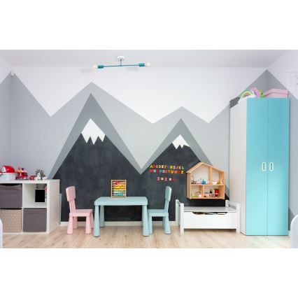 Detský stôl s dvoma stoličkami stojí v izbe s nástennou maľbou hôr. Na stene je abecedná tabuľka s číslicami. V pozadí je detská skriňa.