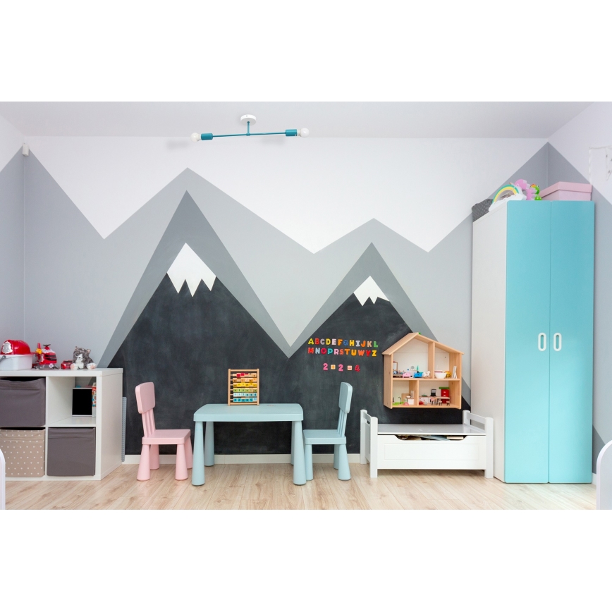 Detský stôl s dvoma stoličkami stojí v izbe s nástennou maľbou hôr. Na stene je abecedná tabuľka s číslicami. V pozadí je detská skriňa.