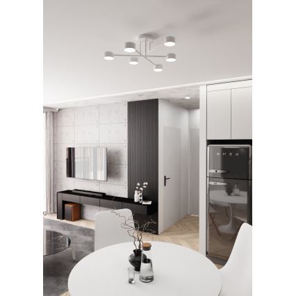 Biela stropná lampa s viacerými svetlami svieti v interiéri. Nachádza sa v jedálenskej časti bytu s bielym stolom, stoličkami a tmavou chladničkou Smeg. V pozadí je obývacia izba s televízorom.