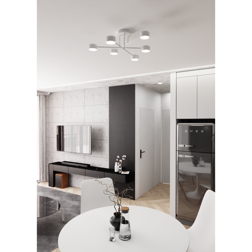 Biela stropná lampa s viacerými svetlami svieti v interiéri. Nachádza sa v jedálenskej časti bytu s bielym stolom, stoličkami a tmavou chladničkou Smeg. V pozadí je obývacia izba s televízorom.
