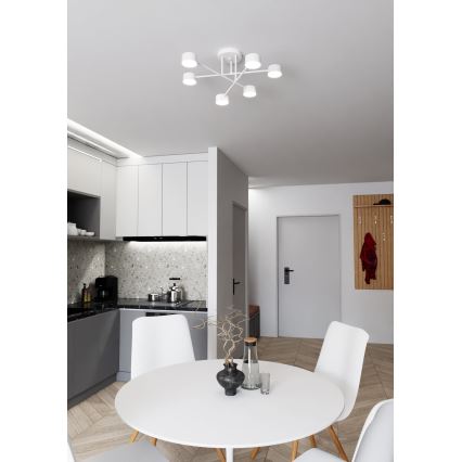 Biela stropná lampa s piatimi svetlami visí v jedálni. Lampa je pripevnená k stropu a jej ramená sa rozvetvujú do strán. Jedálenský stôl s bielou doskou a bielymi stoličkami sa nachádza v priestore s kuchyňou a vstupnou halou.
