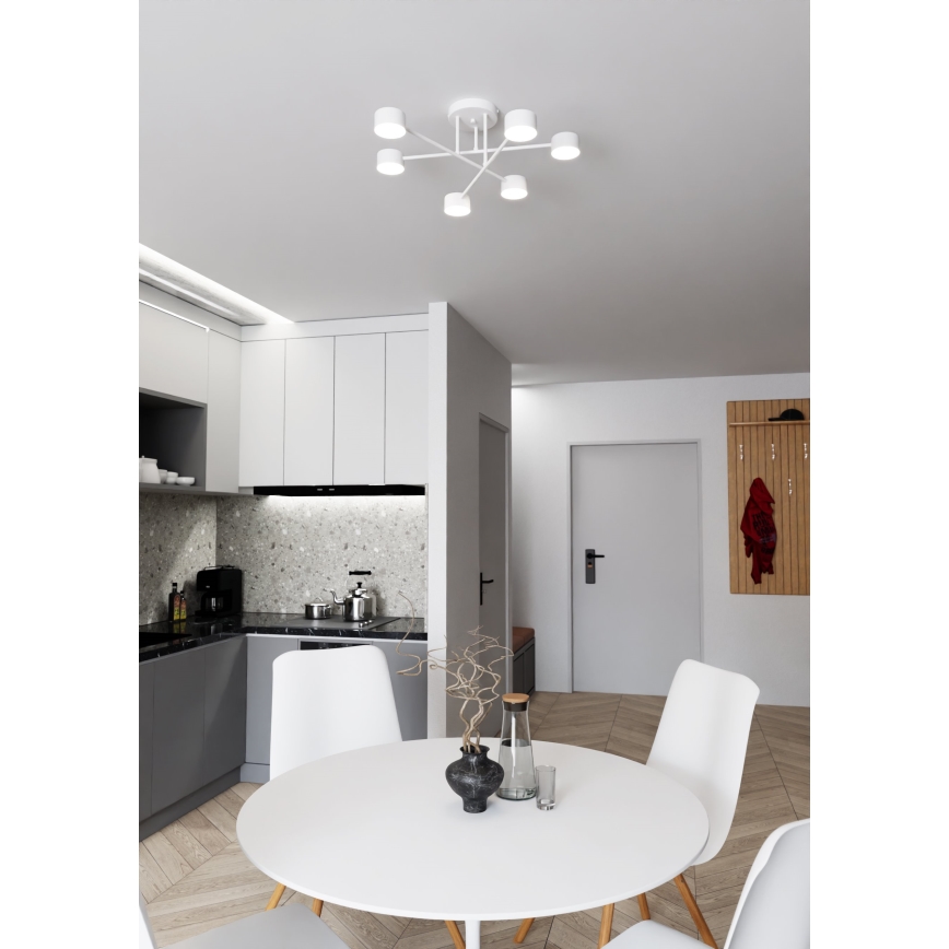 Biela stropná lampa s piatimi svetlami visí v jedálni. Lampa je pripevnená k stropu a jej ramená sa rozvetvujú do strán. Jedálenský stôl s bielou doskou a bielymi stoličkami sa nachádza v priestore s kuchyňou a vstupnou halou.