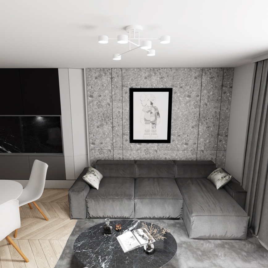 Šedá rohová sedačka stojí v obývacej izbe s betónovou stenou. Na stene visí obraz. Pred sedačkou je nízky čierny stolík. V pozadí je jedálenský kút s bielym stolom a stoličkami.