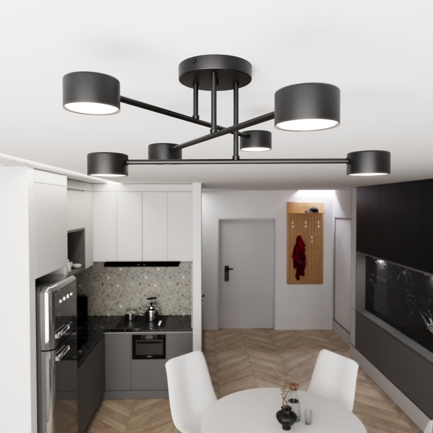 Moderná stropná lampa s piatimi valcovými tienidlami svieti v kuchyni s jedálenským stolom a kuchynskou linkou. Lampa je čiernej farby a má jednoduchý geometrický dizajn. Priestor je zariadený v modernom štýle.