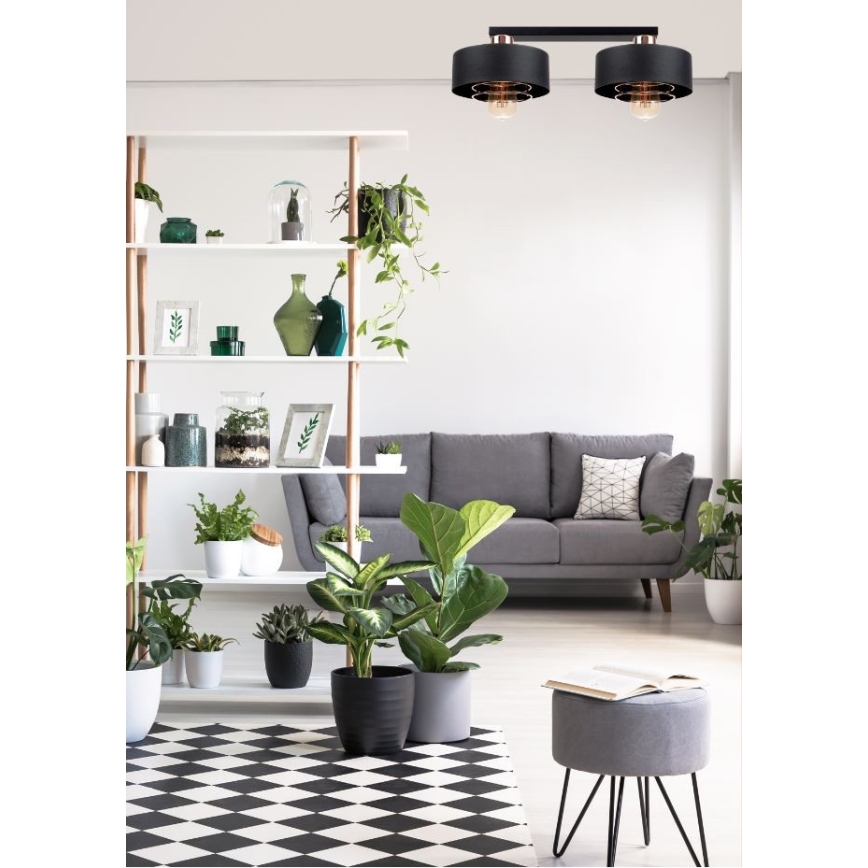 Dvojité stropné svietidlo svieti v obývacej izbe. Svietidlo má čierne tienidlá a medené detaily. Izba obsahuje šedú sedačku, police s izbovými rastlinami a čierno-bielu dlažbu.