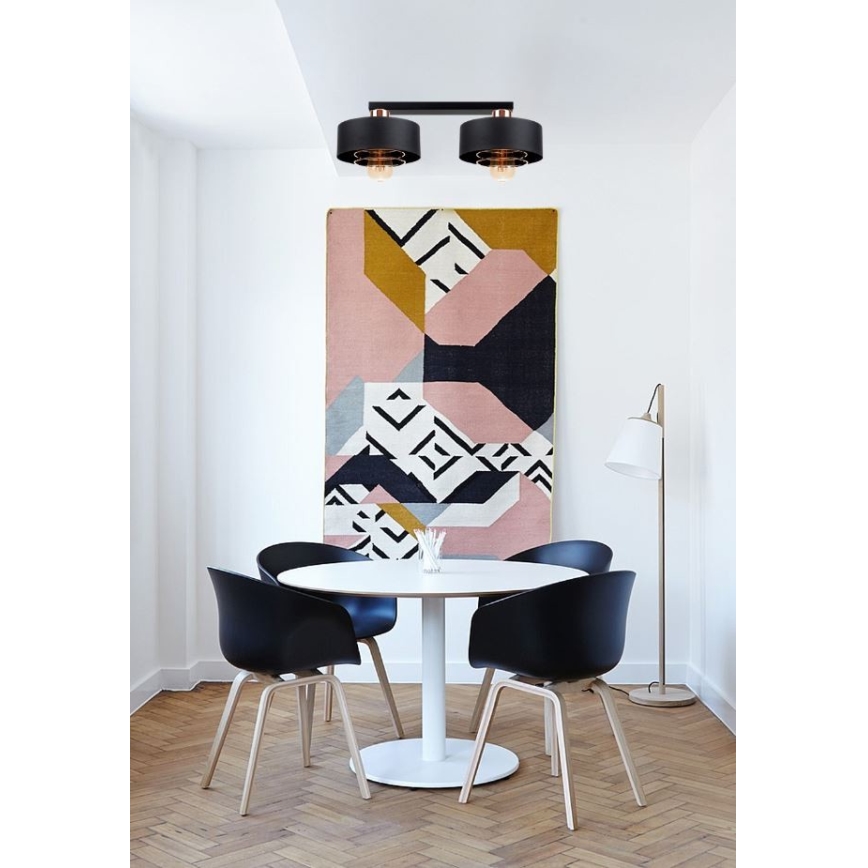 Dvojité stropné svietidlo svieti v jedálni. Svietidlo je čierne s medenými detailmi. V miestnosti sa nachádza jedálenský stôl s čiernymi stoličkami a abstraktný obraz na stene.