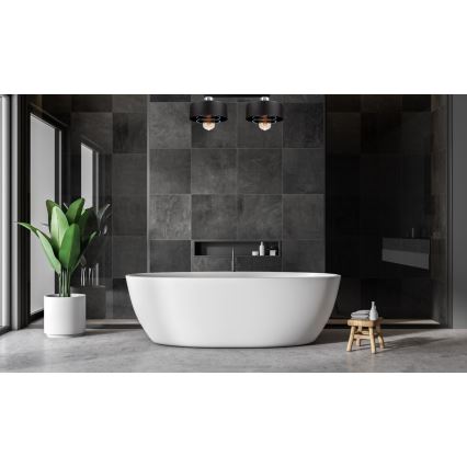 Dve čierne svietidlá svietia v modernej kúpeľni. V strede je voľne stojaca biela vaňa. Steny sú obložené tmavosivými dlaždicami.