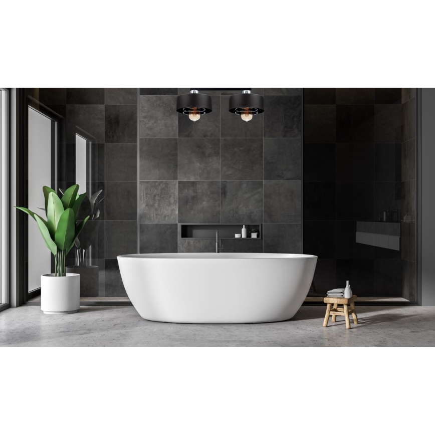 Dve čierne svietidlá svietia v modernej kúpeľni. V strede je voľne stojaca biela vaňa. Steny sú obložené tmavosivými dlaždicami.