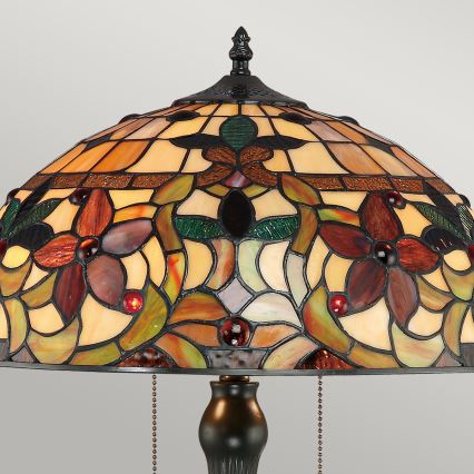 Stolová lampa s tienidlom z vitráže zobrazuje kvetinový motív v hnedých, zelených a béžových tónoch. Tienidlo je polokupolové, s kovovou podstavou. Reťaze visia zo spodnej strany tienidla.