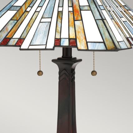 Stolová lampa stojí na jednotnom pozadí. Má tmavú hnedú nohu a kužeľovitý tienidlo z farebného vitráža v odtieňoch modrej, bielej, oranžovej a červenej. Tienidlo je zdobené geometrickými vzormi. Lampa má dva spínače na šnúrke.