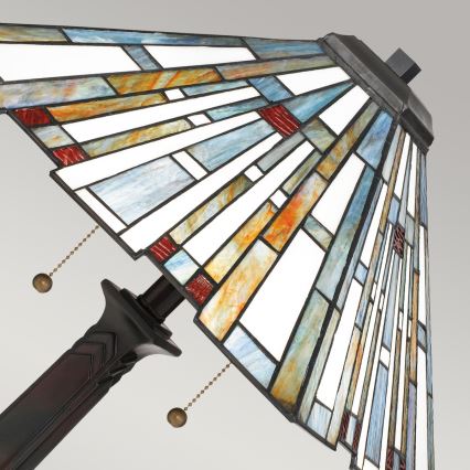 Stolová lampa s tienidlom z farebného skla v geometrickom vzore, v odtieňoch modrej, oranžovej, červenej a bielej, je zobrazená v uhle. Lampa má hnedú kovové nohu s ozdobným prvkom. Na tienidle sú viditeľné retiazky na zapínanie.