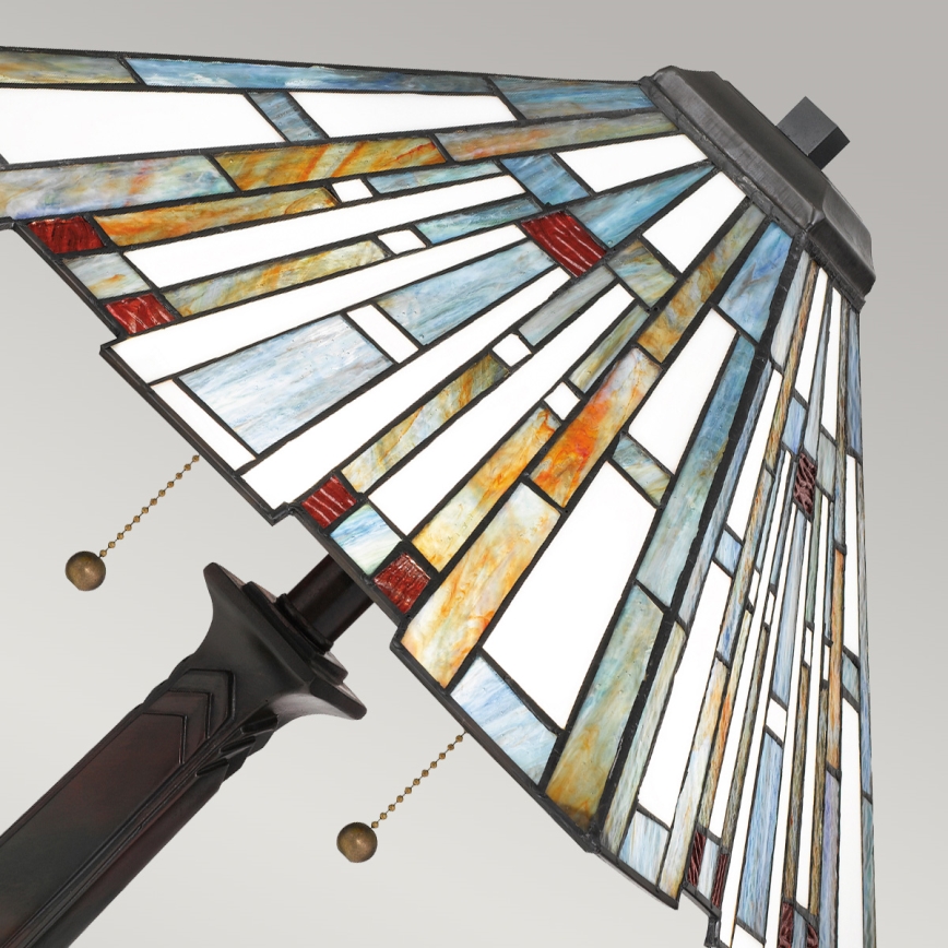 Stolová lampa s tienidlom z farebného skla v geometrickom vzore, v odtieňoch modrej, oranžovej, červenej a bielej, je zobrazená v uhle. Lampa má hnedú kovové nohu s ozdobným prvkom. Na tienidle sú viditeľné retiazky na zapínanie.