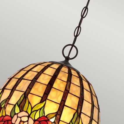 Guľatá Tiffany lampa visí na reťazi. Je zdobená kvetinovými motívmi a štvorcovým vzorom. Lampa je zavesená na svetlom pozadí.