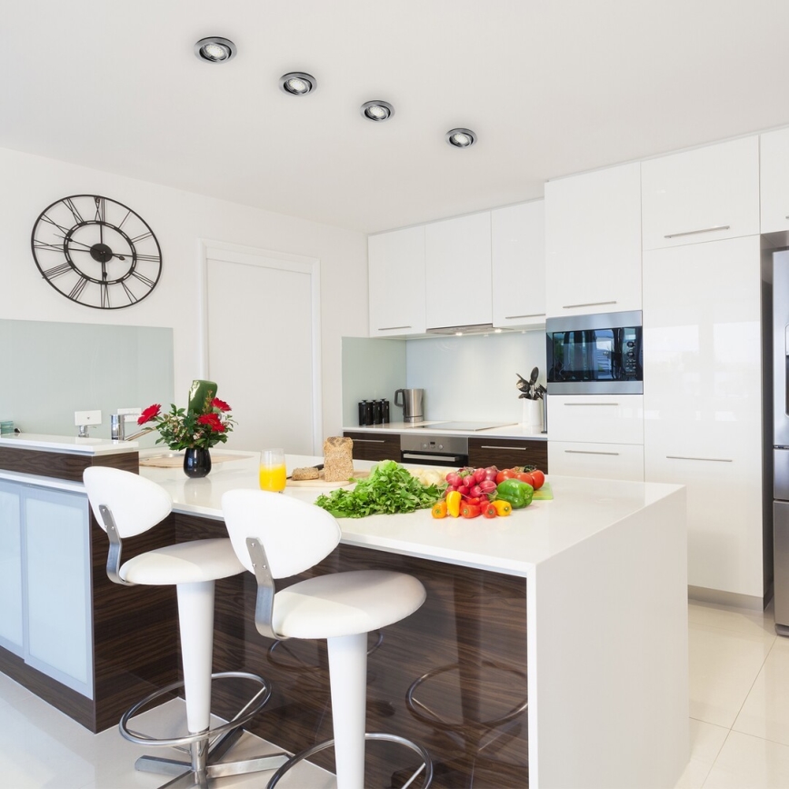 Biela kuchynská linka s ostrovčekom, na ktorom sú umiestnené ovocie a zelenina. Na ostrovčeku sú dve biele barové stoličky. V pozadí je vstavaná mikrovlnná rúra a hodiny na stene.