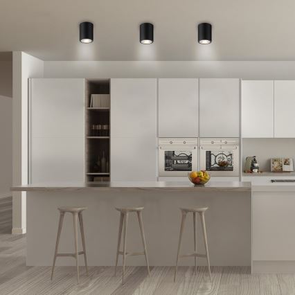 Tri čierne stropné svietidlá svietia v modernej kuchyni s bielymi skrinkami, vstavanými rúrami a drevenou kuchynskou linkou. Na linke stoja tri vysoké stoličky.