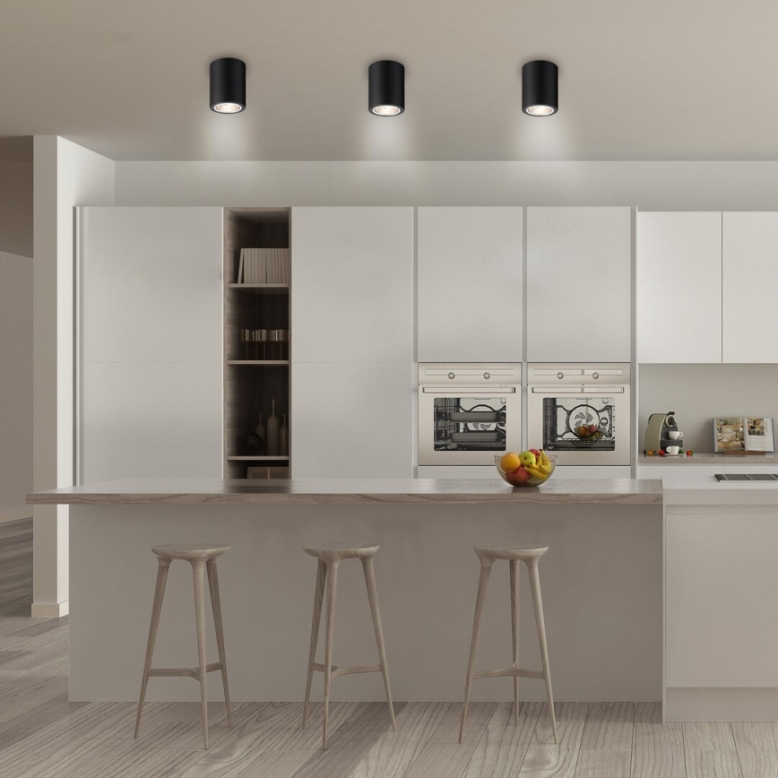 Tri čierne stropné svietidlá svietia v modernej kuchyni s bielymi skrinkami, vstavanými rúrami a drevenou kuchynskou linkou. Na linke stoja tri vysoké stoličky.