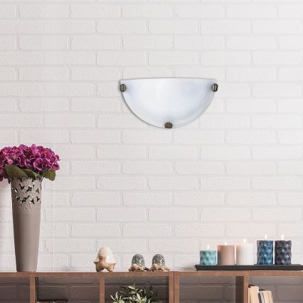 Polovičná nástenná lampa z matného skla je pripevnená na bielej tehlovej stene. Pod lampou sa nachádza drevená polica s kvetináčom, soškami a sviečkami.