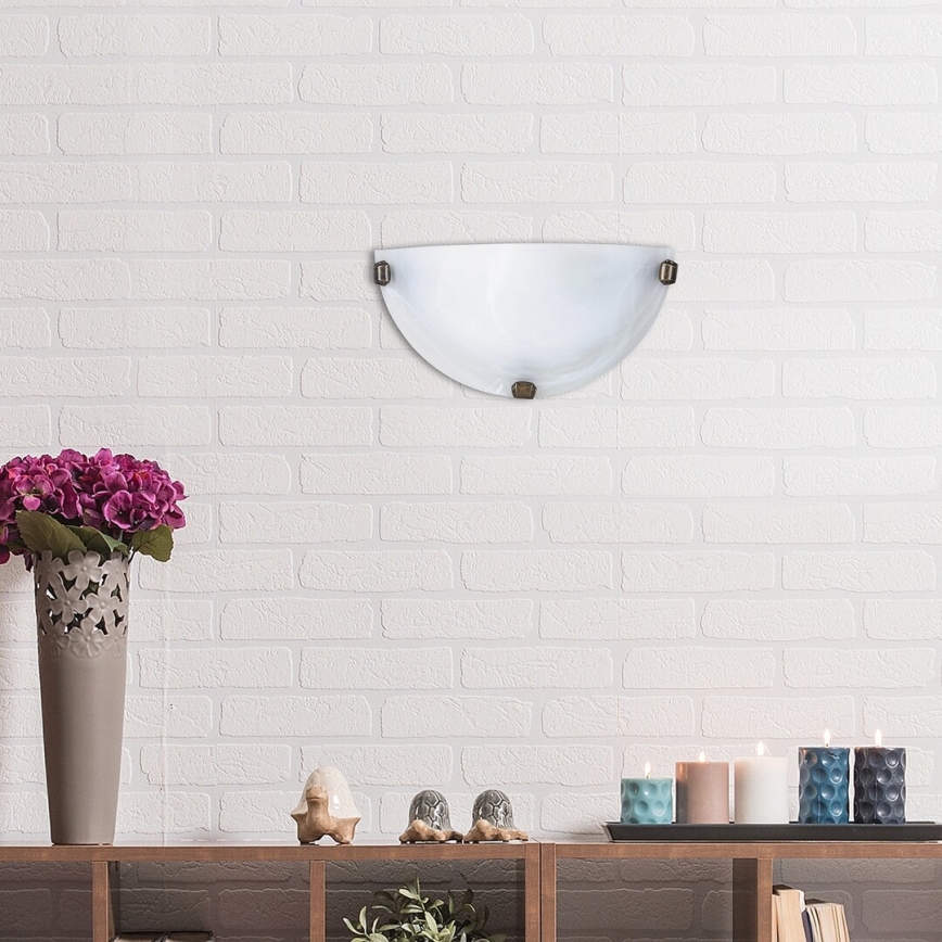 Polovičná nástenná lampa z matného skla je pripevnená na bielej tehlovej stene. Pod lampou sa nachádza drevená polica s kvetináčom, soškami a sviečkami.