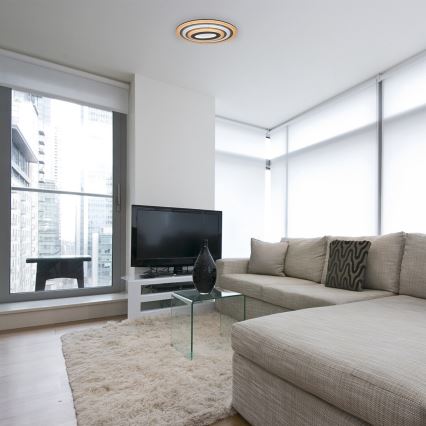 Svetlá stropná lampa svieti v modernej obývacej izbe. Rohová sedačka je umiestnená pred televízorom a nízkym skleneným stolíkom. Okná s výhľadom na mesto sú za stenou.